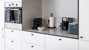 A modern kitchen with white cabinetry, a built-in oven, a coffee maker, and a kettle. - Corner Apartment (Bruxelles)