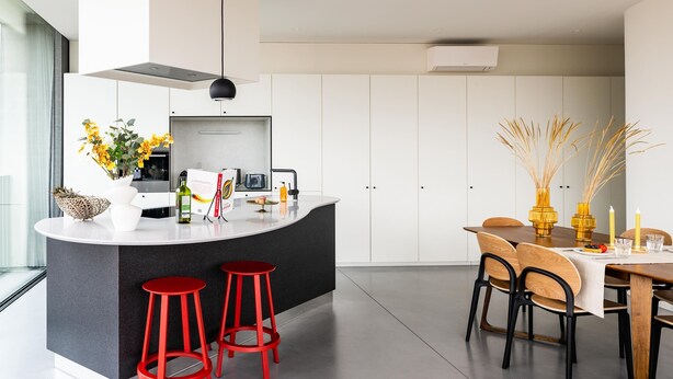A modern kitchen with a central island, red bar stools, and a dining area with a wooden table and chairs. - Corner Apartment (Bruxelles)