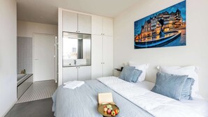 A modern hotel room with a bed, a bedside table with a book and a bowl of macarons, a large window, and a painting of a cityscape. - Corner Apartment (Bruxelles)