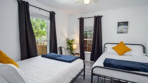 A bedroom with two beds, a ceiling fan, a window with curtains, a small table with a lamp, and a painting on the wall. - Charming 4BR/2BA Only 5 mins Downtown & Riverwalk (San Antonio)