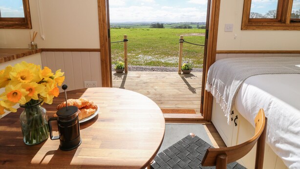 Une pièce chaleureuse avec une table en bois, un lit et une vue sur un champ verdoyant, visible à travers une porte ouverte.