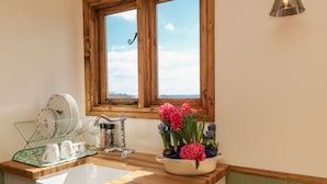 A kitchen with a wooden countertop, a white sink, a window with a view, and a flower vase with pink and purple flowers. - Tawny (Crediton)