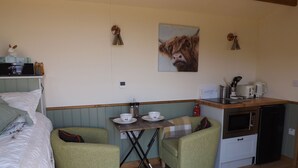 A compact room with a bed, two green armchairs, a small table with cups and a kettle, a microwave, and a painting of a cow on the wall. - Tawny (Crediton)