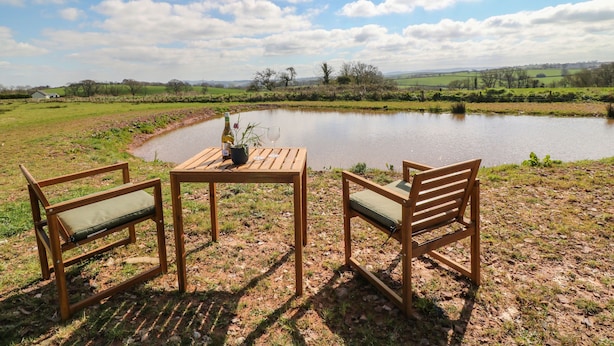 A wooden table with two chairs facing a pond, surrounded by a grassy field with a clear sky. - Tawny (Crediton)