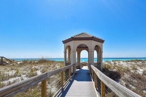 Un paseo marítimo de madera que conduce a un mirador con arcos, con vistas a una playa de arena y vegetación escasa.