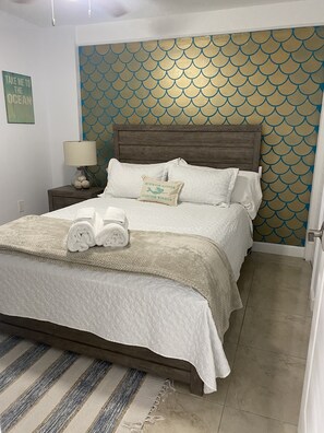 A neatly made bed with a quilted white cover, a wooden headboard, and a decorative pillow with a mermaid and starfish design.