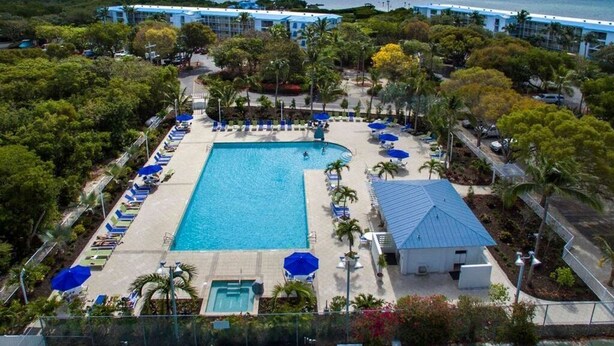 An aerial view of a resort with a central swimming pool, surrounded by lounge chairs and umbrellas, with a backdrop of a beach and ocean. - Ocean Views at Ocean Pointe 
Tavernier, FL (Tavernier)
