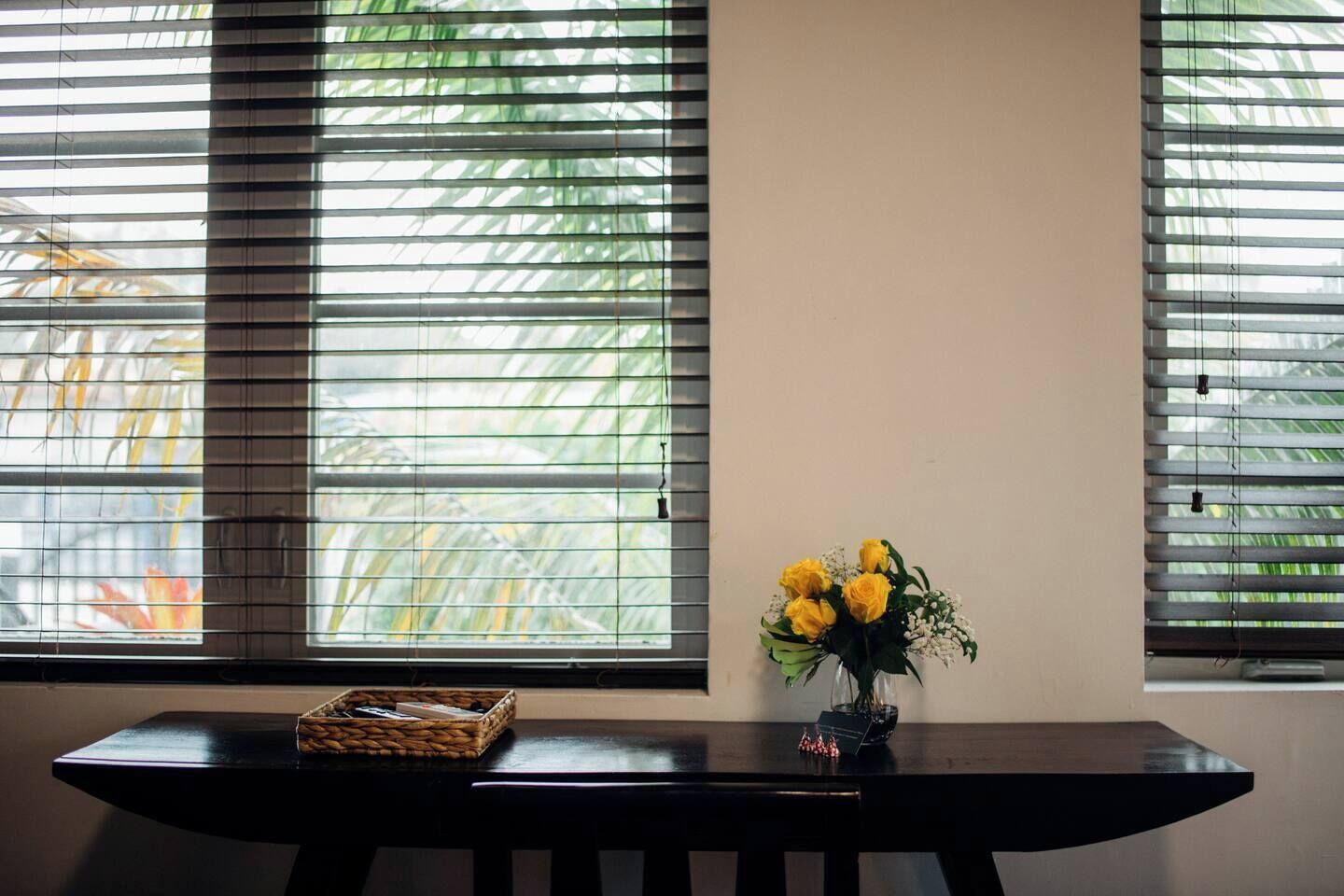 A room with a dark wooden table, a vase with yellow flowers, and a window with blinds.