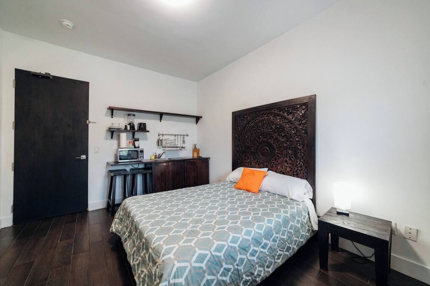 A bedroom with a bed, a headboard, a small table, a nightstand, a kitchen area, and a door.