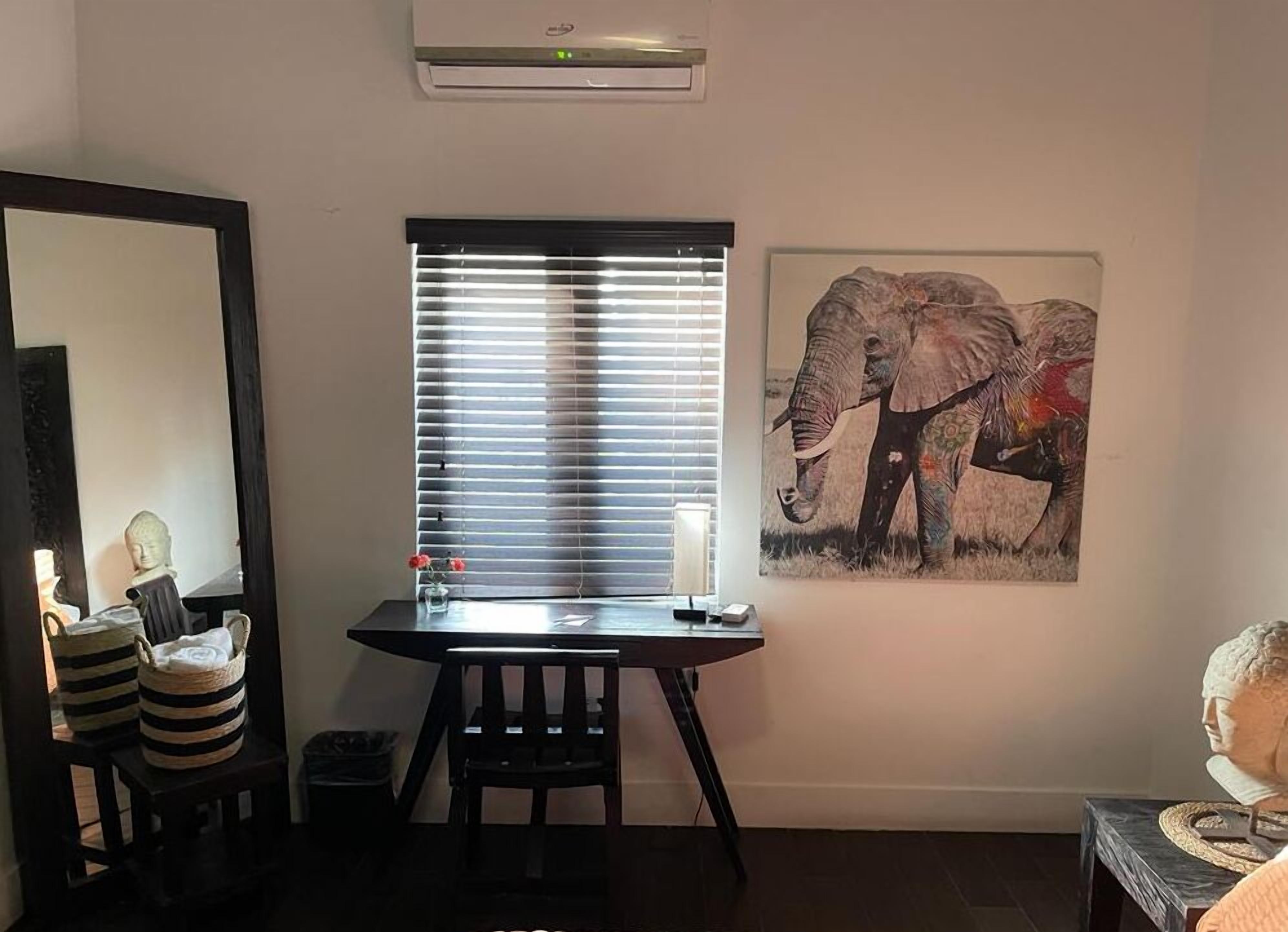 A room with a window with blinds, a painting of an elephant, a bust on a table, and a chair.