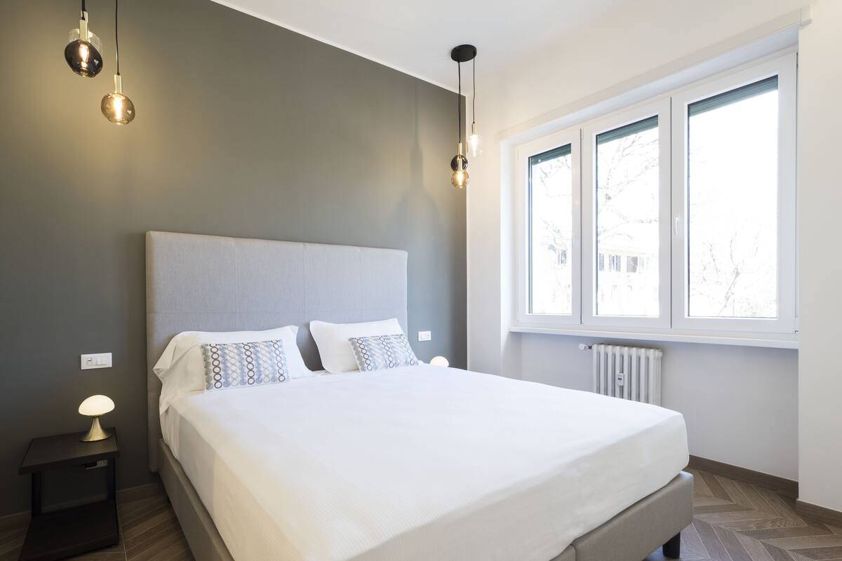 A modern bedroom with a large bed, a nightstand, a lamp, and a window with a view of trees.