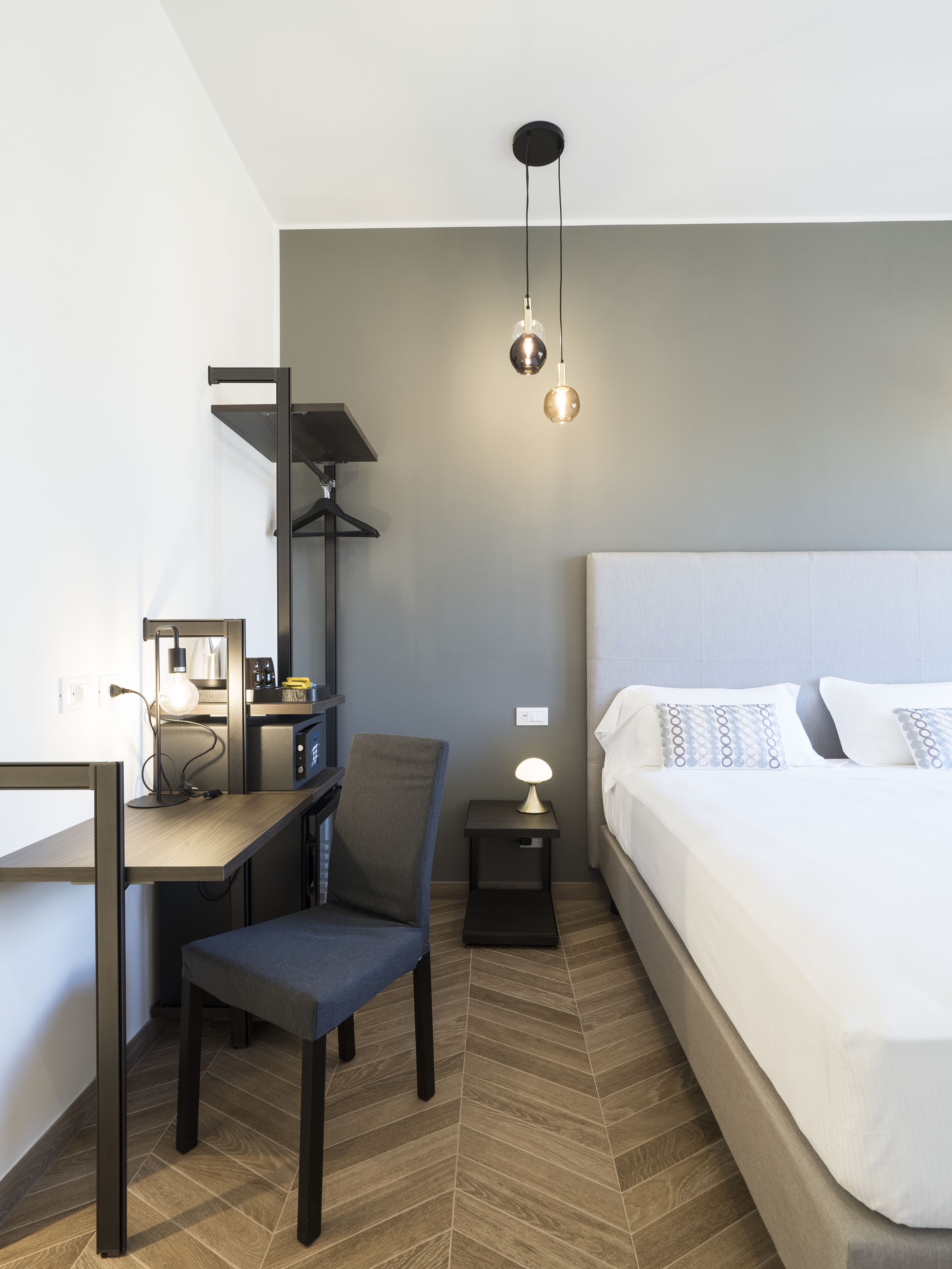 A modern hotel room with a bed, a desk, a chair, and a nightstand with a lamp.