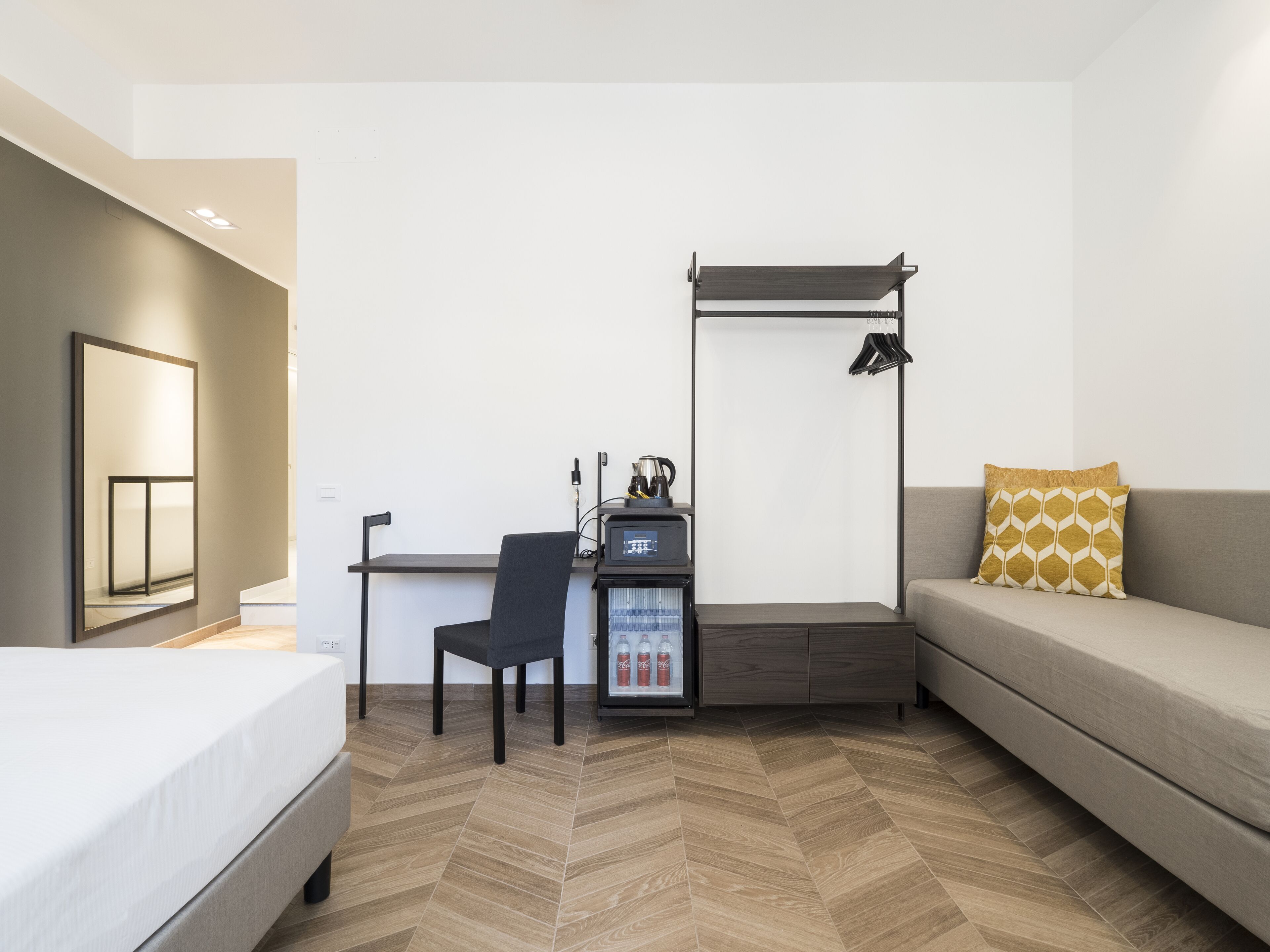 A modern hotel room with a bed, a desk with a computer, a chair, a sofa, and a minibar.