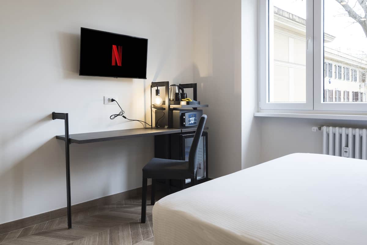 A modern hotel room with a bed, a desk with a computer, a TV mounted on the wall, and a window with a view of buildings.