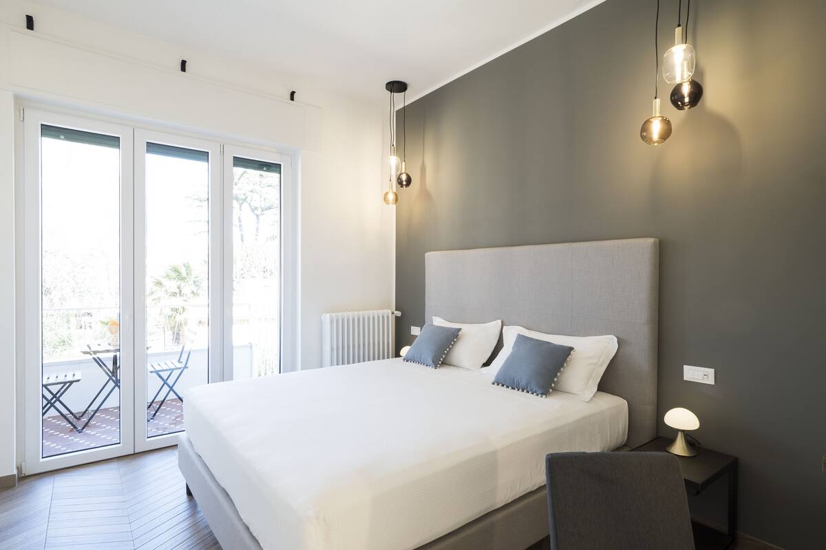 A modern bedroom with a large bed, a nightstand with a lamp, and a view through a sliding glass door to an outdoor patio.