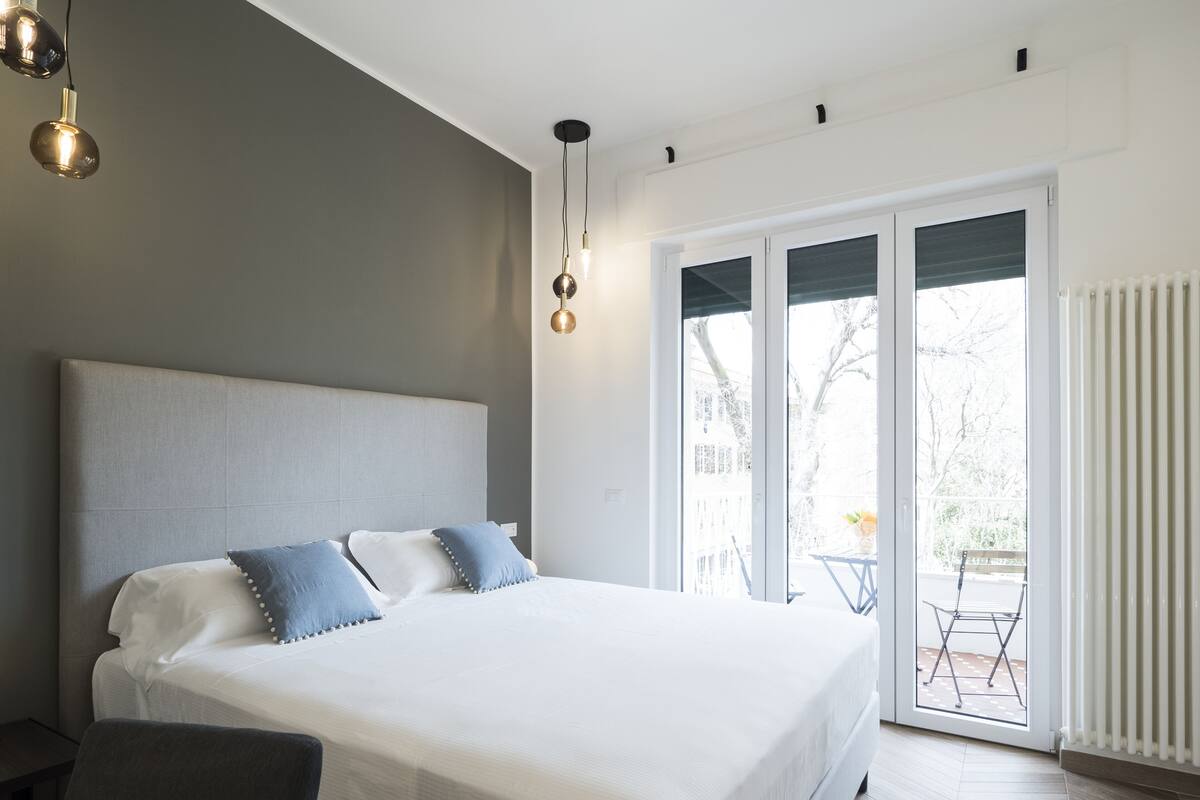 A modern bedroom with a large bed, a desk, and a view of the outdoors through a sliding glass door.