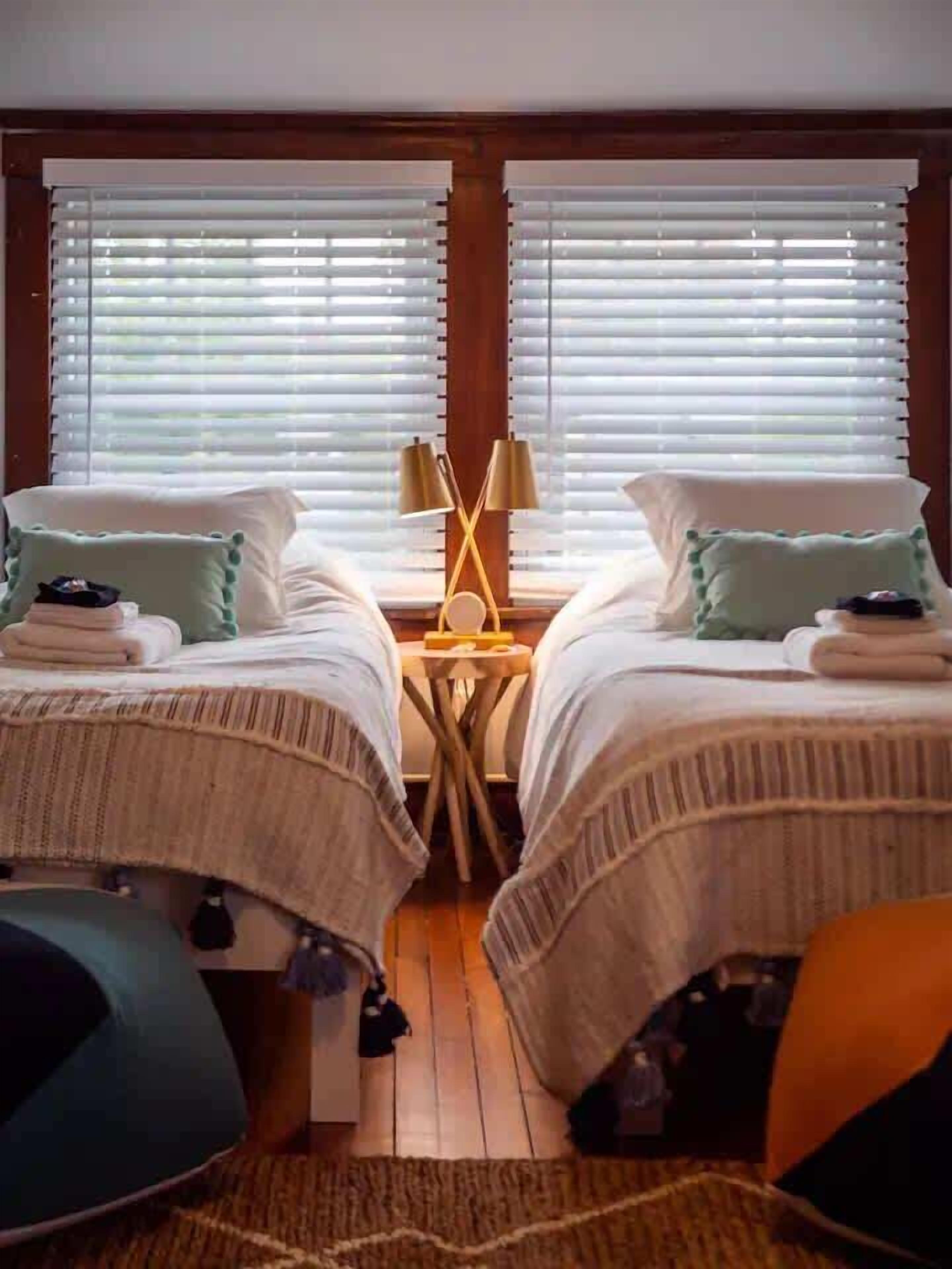 Een tweepersoonskamer met twee bedden, een klein tafeltje met een lamp en uitzicht door de jaloezieën.