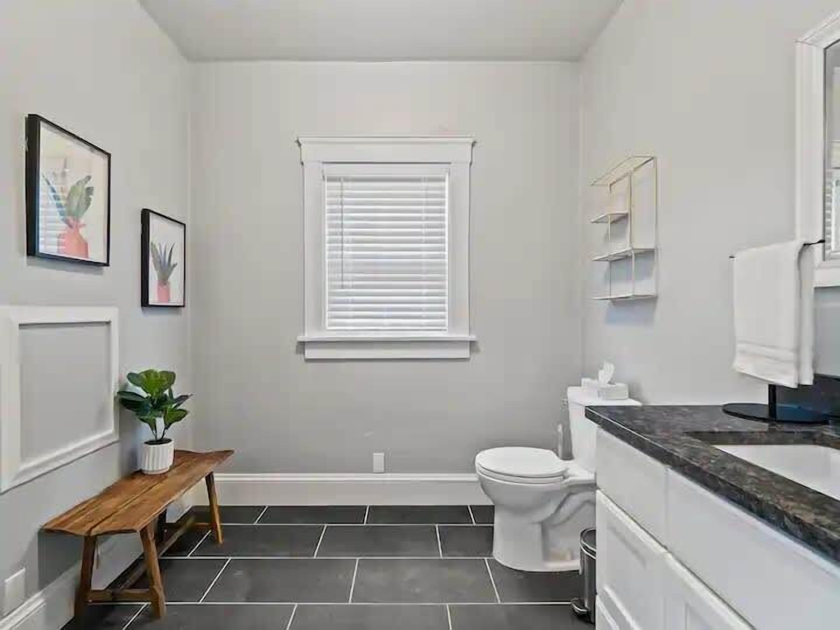 Ein kleines Badezimmer mit Toilette, Waschbecken, Fenster mit Jalousien, einer Sitzbank und einer Topfpflanze.