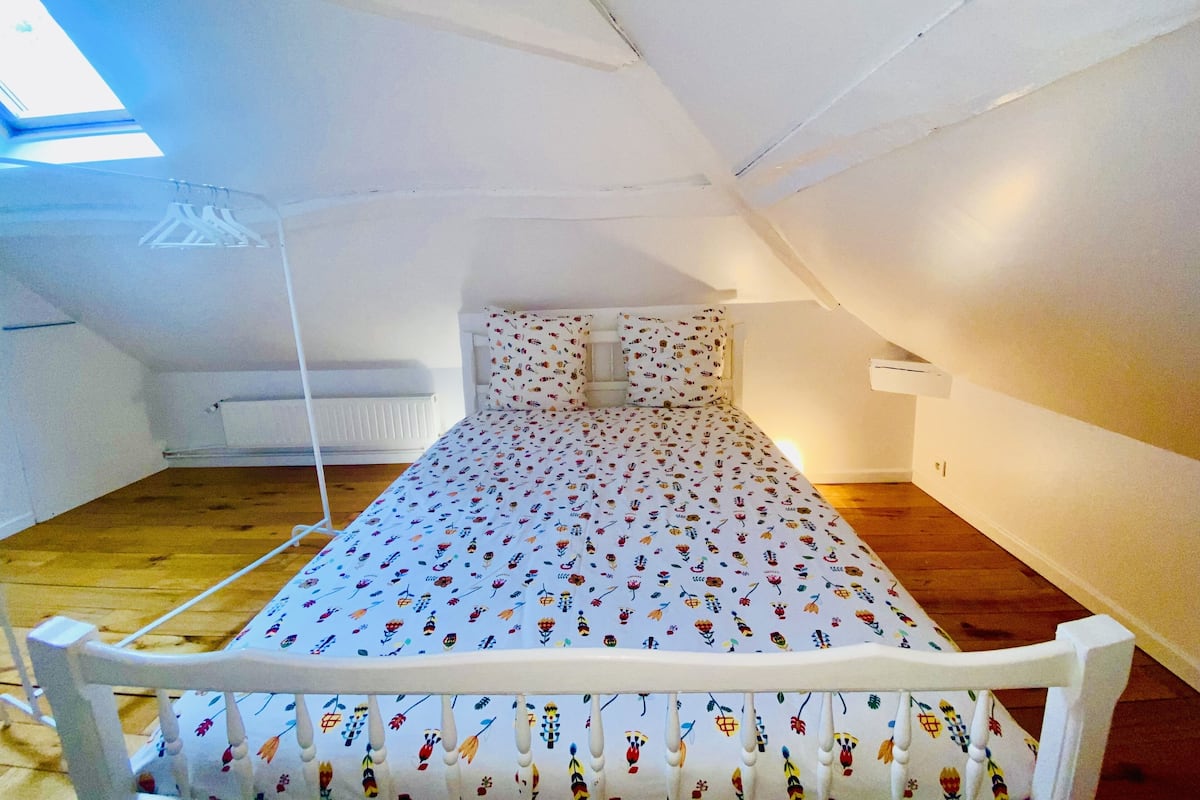 Quarto pequeno com uma cama de solteiro, teto inclinado, paredes brancas e piso de madeira.