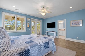 Une chambre à coucher avec un lit, un ventilateur de plafond, une télévision et une salle de bain visible à travers une porte ouverte.