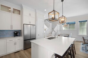 Une cuisine moderne avec des meubles blancs, un réfrigérateur en acier inoxydable, un grand îlot central doté d’un évier et deux luminaires suspendus.