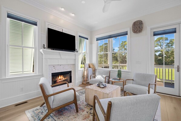 A living room with a fireplace, comfortable seating, a flat-screen TV, and large windows offering a view of the outdoors. - Bonito Bliss - Golf Cart Included! 722 Bonito (Fripp Island)