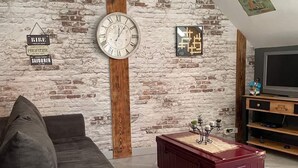 Una sala de estar con pared de ladrillo, un pilar de madera, un reloj, un televisor y una maleta roja.