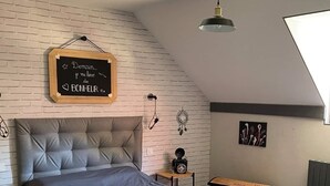 Un dormitorio con techo inclinado, una cama con cabecera gris, una manta roja, una pequeña mesa de madera, un reloj negro y un atrapasueños en la pared.