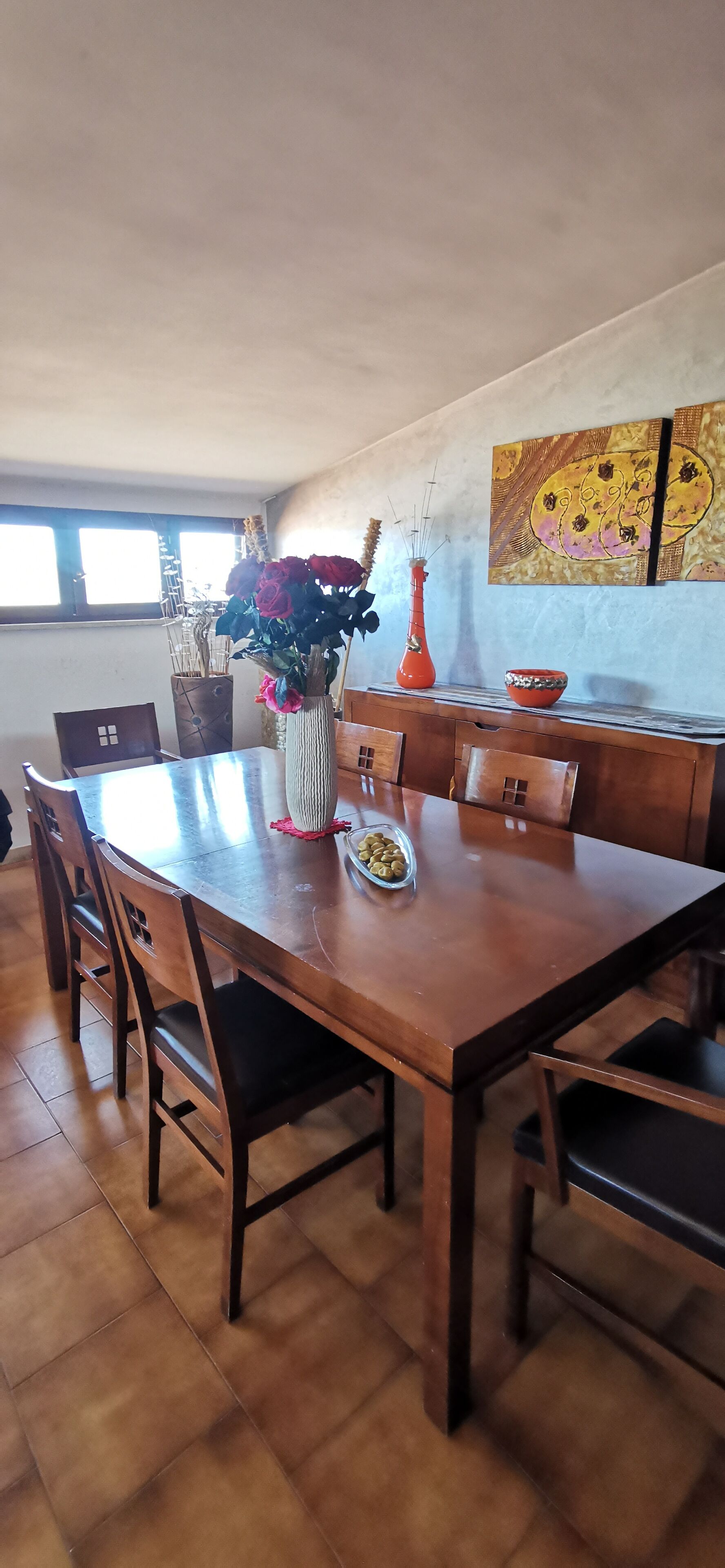 A dining area with a wooden table, chairs, a vase with flowers, and a painting on the wall.