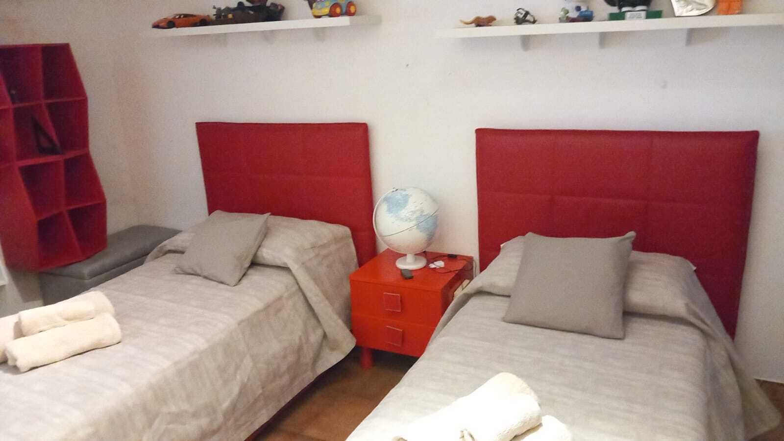 Habitación doble con dos camas, cada una con cabecero rojo, una mesita de noche pequeña con un globo terráqueo y un estante sobre las camas.
