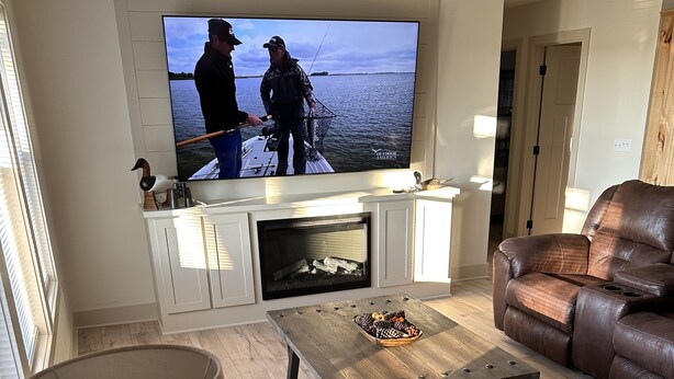 A living room with a fireplace, leather sofa, and a television displaying a fishing scene. - Outdoorsman Paradise (Wolf Lake)