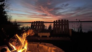 Una fogata con troncos y brasas ardientes, una silla Adirondack de madera, y una vista del atardecer sobre un cuerpo de agua.
