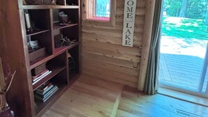 Interior de una cabaña de madera con una estantería, un ventanal pequeño y un cartel que dice 'HOGAR, DULCE LAGO'.