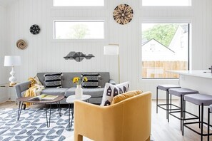 Ein modernes Wohnzimmer mit einem grauen Sofa, einem gelben Sessel und Barhocker. An den Wänden sind dekorative Wandbilder angebracht und durch ein Fenster ist ein Haus zu sehen.