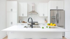 A modern kitchen with white cabinetry, a central island with a sink, a stainless steel range hood, and a refrigerator. - Dapper Dan (Atlanta)