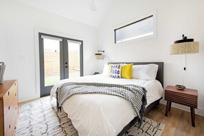 Quarto com uma cama, mesa de cabeceira de madeira, abajur e vista pela porta de correr.