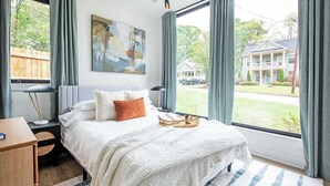 A bedroom with a large window, a bed with white bedding, a bedside table with a lamp, and a painting on the wall. - Dapper Dan (Atlanta)
