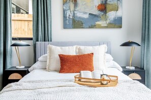 A neatly arranged bedroom with a large bed, two bedside tables with lamps, a framed abstract painting, and a window with curtains. - Dapper Dan (Atlanta)