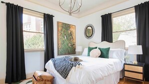 A bedroom with a large bed, a bedside table with a lamp, a window with curtains, a painting on the wall, and a small rug.