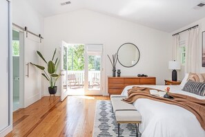 A bedroom with a bed, bedside table, dresser, and a large mirror. There is a window with a view of greenery outside.