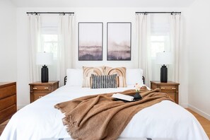 Makuuhuoneessa on suuri sänky, kaksi yöpöytää, yölamppu, ikkuna, jossa on verhot, ja kaksi seinälle kiinnitettyä valokuvakehystä.