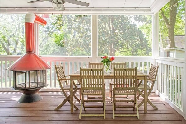 Une terrasse en bois avec une table et des chaises, un chauffage d’extérieur rouge et une vue sur les arbres à travers de grandes fenêtres.