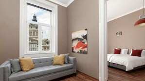 A room with a grey sofa, a bed with red and white bedding, a large window with a view of historic buildings, and a red pendant light. - Venus Apartments - Italian Stylish (Budapest)