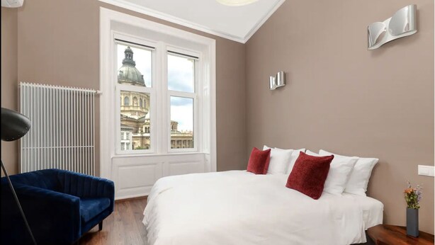 A bedroom with a large bed, a blue chair, a radiator, and a view of a building through the window. - Venus Apartments - Italian Stylish (Budapest)