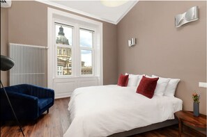 Una camera da letto con un letto grande, una poltrona blu, un termosifone e vista su un edificio dalla finestra.