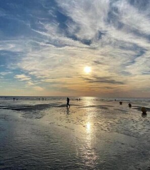 A beach with people walking, the sun setting, and a clear sky.