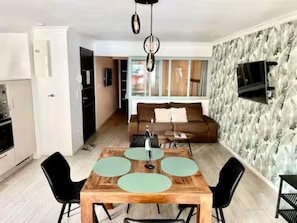 A modern living room with a wooden dining table, black chairs, a brown sofa, and a TV mounted on a textured wall. - Rêves Tranquille 50 metres from the beach and town center (Mers-les-Bains)