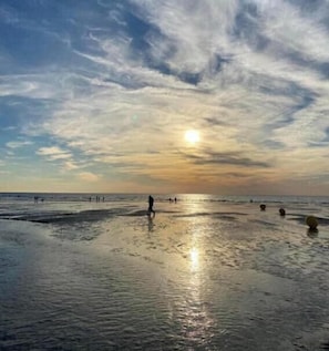 A beach with people walking, the sun setting, and a clear sky.