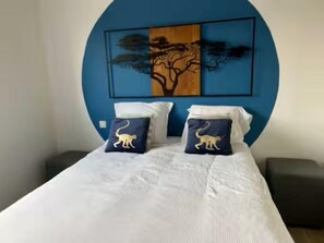 Cama com roupa de cama branca, duas almofadas com estampas de animais e parede azul com pintura de árvore.