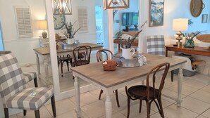 A dining area with a wooden table, chairs, and a chandelier. There is a TV in the background and a painting on the wall. - “Cottage-Like” 2 Beds/2 Bath Condo Close To Beaches & The Heart Of ‘Old Stuart' (Stuart)